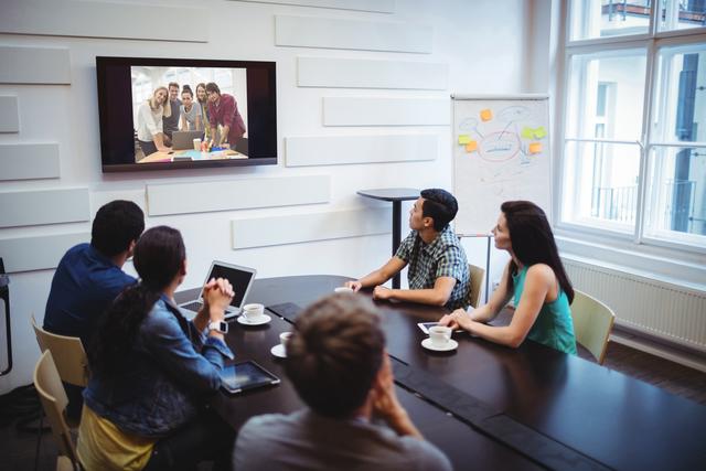 Business executives are engaged in a video conference in a modern conference room. They are seated around a table, focusing on a screen displaying a group of colleagues. This image is ideal for illustrating concepts related to remote work, corporate meetings, teamwork, and modern office environments. It can be used in business presentations, corporate websites, and articles about professional collaboration and technology in the workplace.