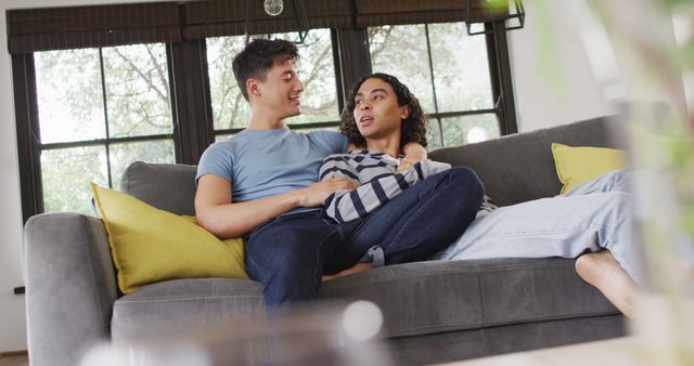 Young interracial couple sits comfortably on a gray sofa in a well-lit living room, engaging in a relaxed conversation. The setting includes large windows that allow natural light to flood the space, giving a cozy and inviting atmosphere. Ideal for depicting themes of domesticity, relationships, modern living, and relaxation in a home environment.
