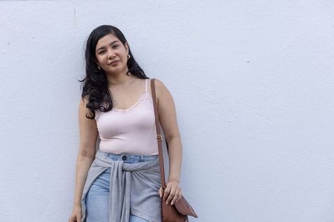 Young woman leaning on pale wall holding brown leather bag wearing casual pastel outfit