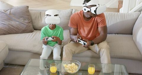 Father and Son Playing VR Games on Sofa Wearing White VR Headsets and Using Controllers