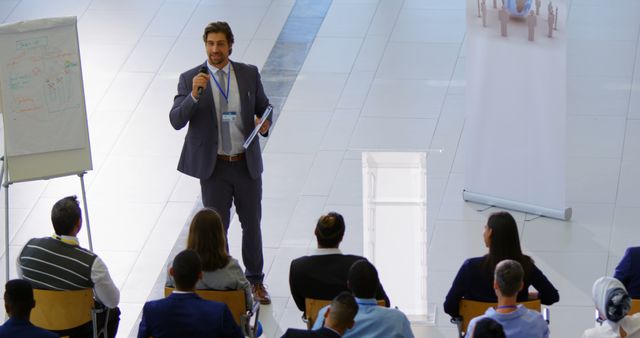 Businessman presenting to diverse group of professionals seated in a modern conference venue. Ideal for illustrating corporate events, leadership training, business strategy discussions, professional presentations, and public speaking engagements.