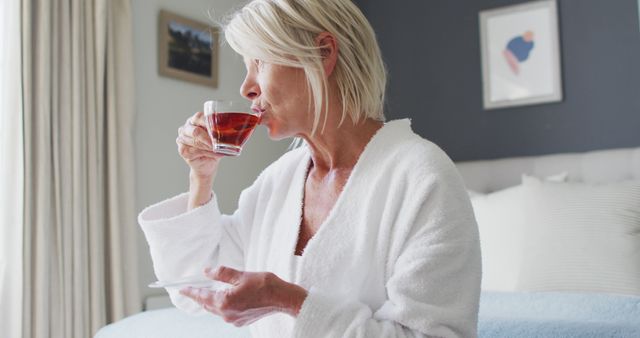 Senior woman enjoying a moment of relaxation while sipping tea at home in a white robe. This image can be used to depict themes of self-care, healthy lifestyle, and peaceful morning routines. Ideal for articles, adverts, or promotions centered on health, wellness, and senior living.
