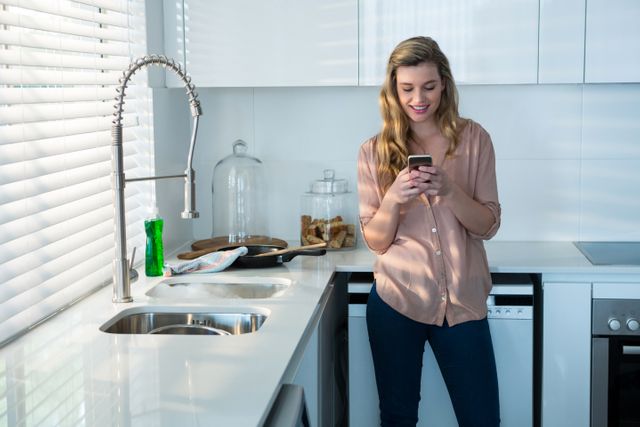 The image shows a young woman standing in a modern kitchen, using her smartphone. She appears relaxed and casually dressed, reflecting a typical at-home environment. This image can be used for lifestyle blogs, technology ads, home decor websites, and articles about home life, cooking, or modern living.