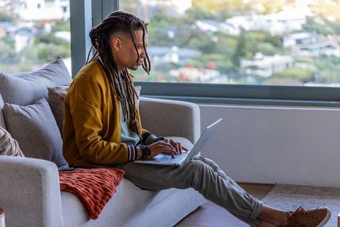 African american man working remotely on laptop while sitting on modern sofa by window