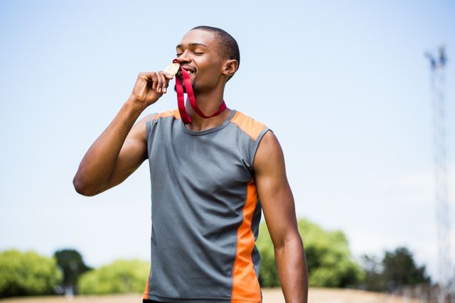 Athlete celebrating victory by kissing his gold medal in an outdoor stadium. Ideal for use in sports-related content, motivational materials, fitness promotions, and articles on athletic achievements and success stories.