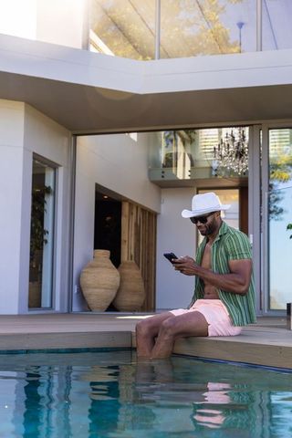 African american man sitting by pool checking smartphone at modern luxury villa terrace
