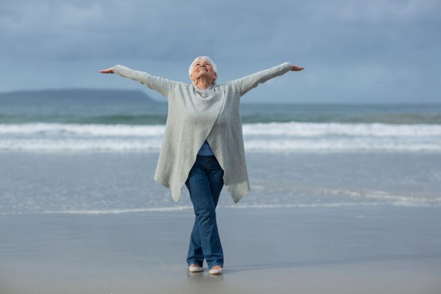 This image depicts a joyful senior woman on the beach with her arms wide open, enjoying the ocean and outdoor environment. It is ideal for use in advertisements, articles, and promotional materials related to healthy aging, outdoor activities, senior wellness, retirement, and the benefits of spending time in nature.
