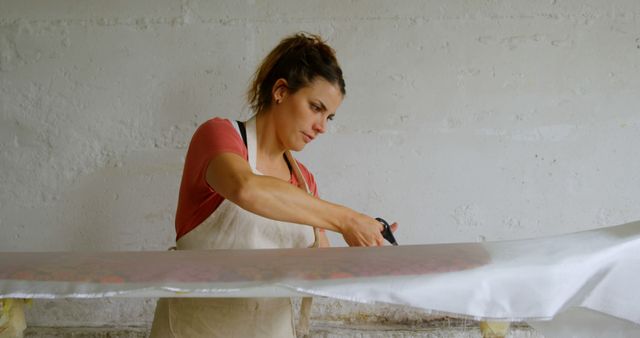 Woman focused on trimming the edges of a large art canvas in a studio setting. Perfect for use in websites or materials related to creativity, artwork, crafting, DIY projects, and studio environments.