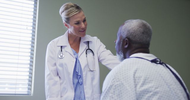 Doctor in white coat with stethoscope around neck consulting with elderly male patient in hospital gown. Window with blinds lights sunny with green wall in the background. Image suitable for healthcare websites, medical articles, hospital promotions, patient-doctor interactions, and health care service brochures.