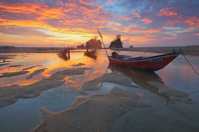Traditional fishing boats moored on a beach with calm waters and a vibrant sunset creating a stunning reflection in the water. Coastal landscape is serene and peaceful with vividly colored skies and gentle waves. Ideal for use in travel brochures, nature and landscape photography collections, beach-themed marketing materials, and inspirational wall art.