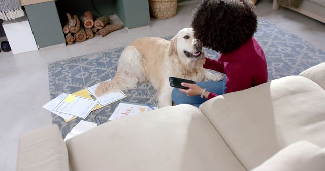 A woman is sitting on the floor in a cozy living room with documents and a smartphone, interacting with her dog. This image can be used to depict remote work, work-life balance, or the bond between pets and their owners. Suitable for articles and advertisements related to home office setups, pet-friendly environments, or lifestyle blogs focused on modern living.