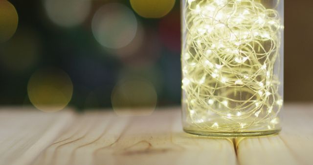 Fairy lights in a glass jar create festive and warm lighting on a wooden table. Great for holiday season décor, event planning, home ambiance, or party invitations. Bokeh effect in background adds warm, cozy feel.
