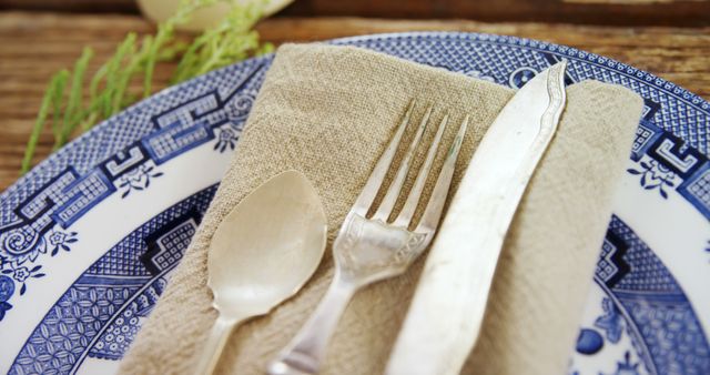 Close-up of a rustic table setting featuring vintage cutlery including a fork, knife, and spoon placed on a folded brown napkin on top of a blue-patterned plate. Ideal for content related to dining, rustic decor, and vintage dining sets.