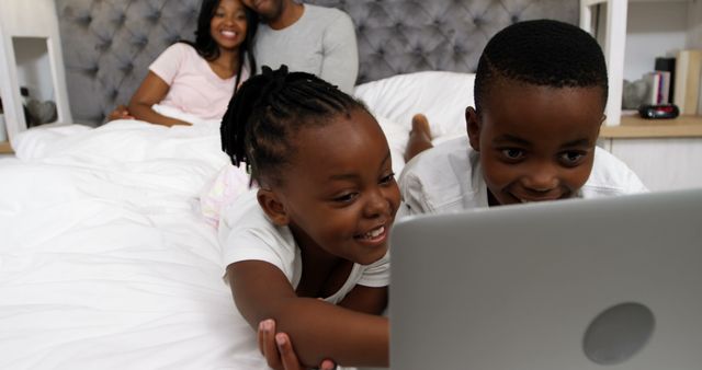 This image shows an African American family relaxing in a cozy bed while the children are using a laptop. The parents are smiling in the background, while the children are engaged with the laptop. Could be used for themes like family bonding, home lifestyle, technology use, parental care, and togetherness.