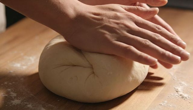 Pressing hands shaping round bread dough on wooden board with scattered flour
