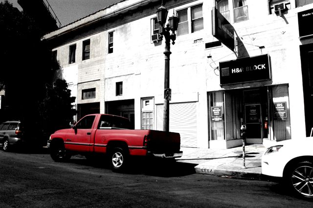 This image captures a striking red pickup truck parked on a black-and-white urban street, highlighting the contrasting colors. The scene includes an old building with architecture reflecting a historical area and a street lamp. This image can be used to represent city life, vintage automobiles, or historical urban settings in advertisements, blog posts, or digital designs.