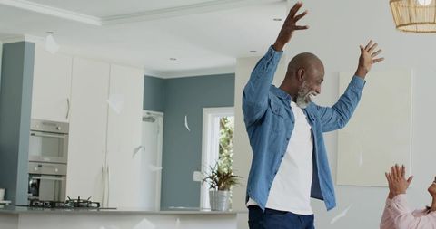 Diverse couple celebrating in modern kitchen, man raising arms, partner clapping with papers