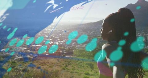 Woman standing on hillside wearing earbuds facing city with augmented reality overlays