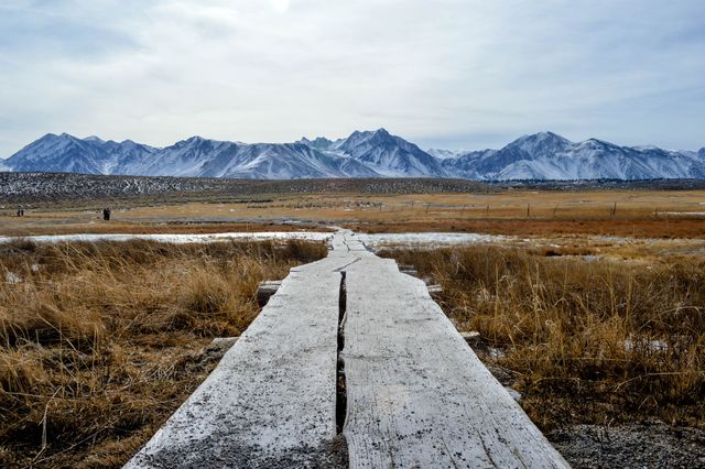 Image shows a rustic wooden path stretching into a vast plain, which leads to snow-capped mountains in the background. Ideal for travel magazines, adventure blogs, outdoor lifestyle promotions, and website hero images showcasing natural beauty and serenity.