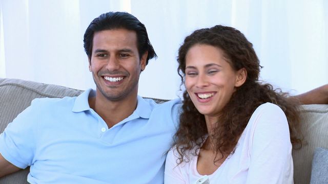 A happy couple is sitting on a comfortable sofa at home, watching television and smiling. They are in a casual and cozy environment, suggesting a moment of relaxation and leisure. This video can be used for promoting home furnishings, family lifestyle, and entertainment products. It can also suit articles about relationships, happiness, and quality family time.