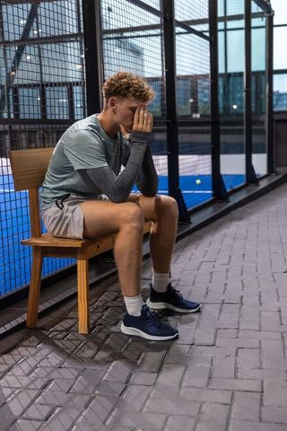 Young athlete sitting distressed on bench beside rooftop paddle court at dusk