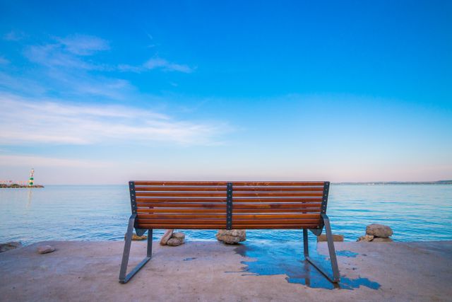 Empty bench facing peaceful ocean under clear blue sky, evokes solitude and calmness. Perfect for concepts of relaxation, tranquility, and solace, suitable for backgrounds, meditative content, and nature promotion.