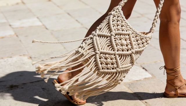 Walking woman wearing tan sandals and cream macrame fringe bag swinging on sunlit pavement