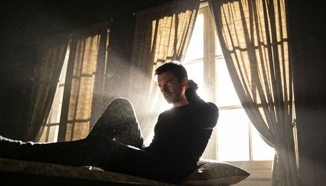 Moody backlit portrait contemplative man sitting on bed with sunbeams dust motes light