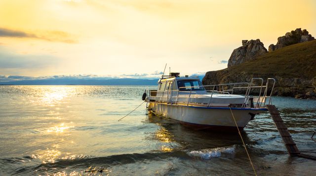 This photo showcases a boat anchored peacefully in a tranquil bay as the sun sets, emitting a golden glow onto the water. Perfect for travel blogs, vacation promotions, nature documentaries, and websites focusing on relaxation and seaside adventures. It can also be used for inspirational and motivational content related to serenity and nature's beauty.