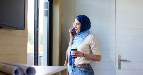 Indian professional woman in blue hijab standing by window holding coffee speaking on phone