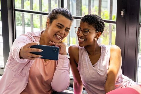 African american friends smiling and taking selfie in pink activewear by sunlit window