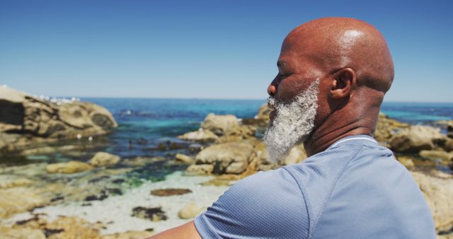 Elderly man with gray beard sitting peacefully by coastal rocks and clear ocean under a bright blue sky. This image can be effectively used in projects themed around meditation, mindfulness, senior wellness, nature retreats, and holistic health promotions.