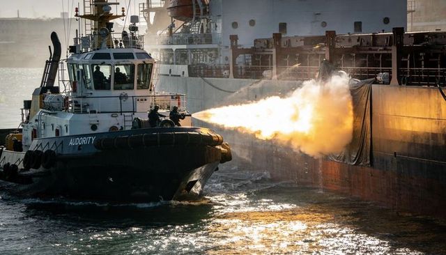 Tugboat firing water cannon at cargo ship hull during firefighting drill at golden hour