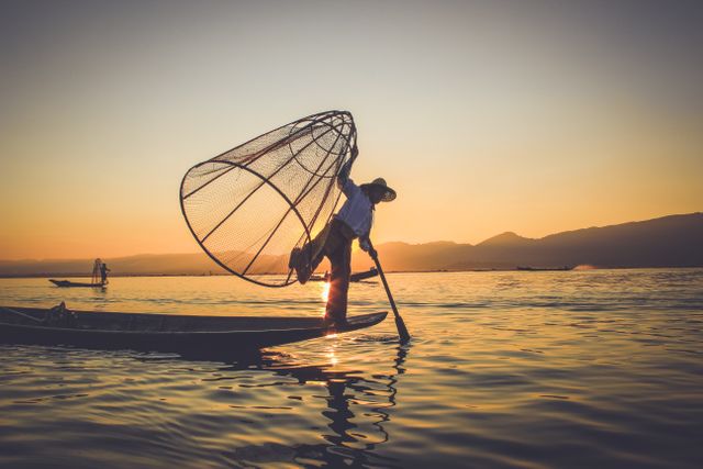 Fisherman is seen balancing on the edge of a canoe while holding a large fishing net at sunrise. The serene ocean and mountainous background make it a visually striking scene. This stock photo can be used for topics related to traditional fishing, rural culture, nature, and the beauty of sunrise moments.