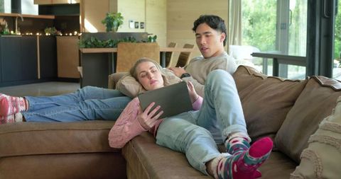 Young couple relaxing on sectional sofa using tablet in modern cozy open-plan living room