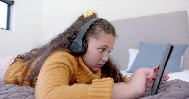 Girl wears headphones while using a digital tablet, lying on bed in a cozy home setting. Perfect for themes involving childhood, technology, online learning, or home entertainment. Suitable for educational content, tech lifestyle blogs, or home living advertisements.