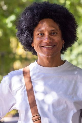 Smiling man with afro standing in sunlit park wearing white t-shirt and crossbody strap with nose ho