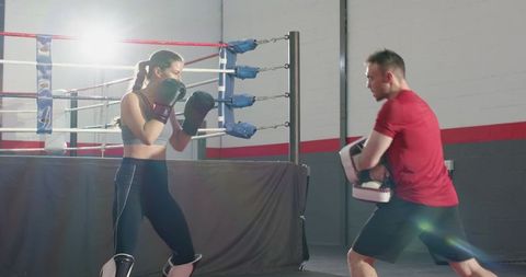 Female boxer training with coach hitting focus pads in ring corner with dramatic lensflare