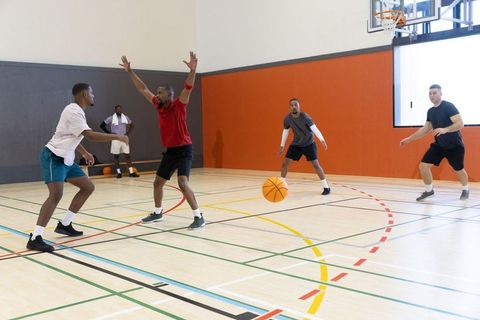 Multiracial men playing pickup basketball on indoor hardwood court, defender raising arms
