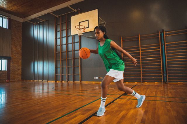 Biracial female basketball player practicing dribbling skills in an indoor court. Ideal for use in sports training materials, fitness and athletic promotions, team sports advertisements, and educational content about basketball techniques.