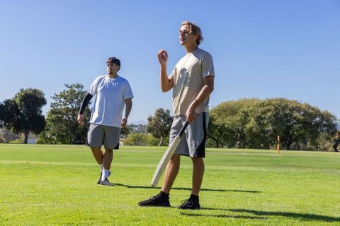 Young men playing casual cricket on sunny park field holding bat and ball during practice