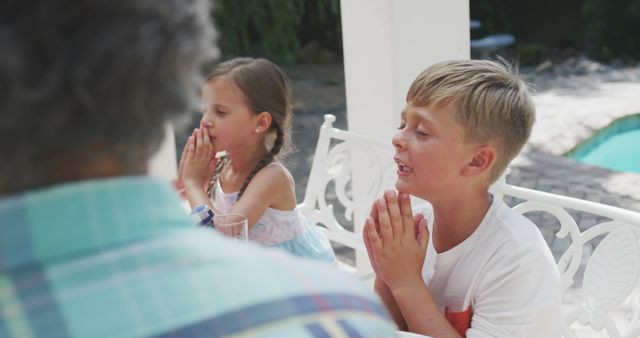 Young boy and girl holding their hands in prayer at an outdoor dinner table. Ideal for use in content about family gatherings, holiday traditions, expressions of faith, acts of gratitude, and kid-friendly activities that teach important values. Can be used in websites, blogs, or articles focusing on family, religion, or inspirational content.