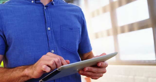 Midsection of caucasian casual businessman in blue shirt using tablet in sunny room, copy space. Communication, creative business, connection and lifestyle, unaltered.