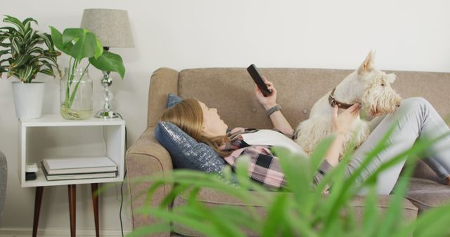 Woman lying on a comfortable couch with a dog, using a smartphone at home. Ideal for articles on leisure, home comfort, pet companionship, and casual living. Useful for promotional materials related to furniture, lifestyle blogs, and pet-friendly environments.