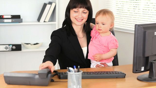 Businesswoman holding baby while working at desk in office. Ideal for illustrating themes of work-life balance, multitasking, parenthood, and modern family dynamics in professional environments. Suitable for articles, blogs, and campaigns focusing on working mothers, career women, and corporate childcare solutions.