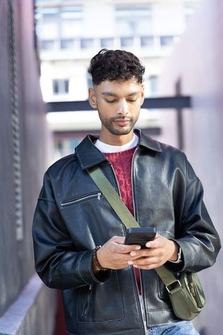Indian man standing in urban corridor using smartphone, wearing leather jacket and crossbody bag