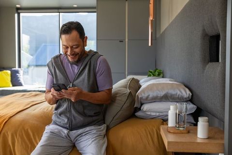 Middle-aged asian man relaxing on bed edge using smartphone in sunlit modern bedroom