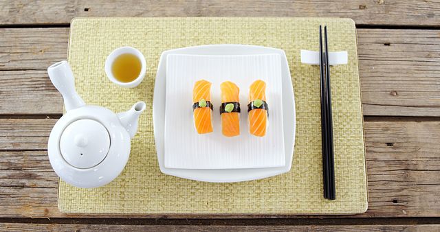 A neatly presented sushi meal with salmon nigiri is accompanied by a cup of tea and chopsticks, with copy space. The simplicity of the arrangement on the wooden surface emphasizes a traditional Japanese culinary experience.