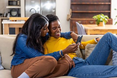 Laughing female partners sharing smartphone while lounging on sofa in cozy living room
