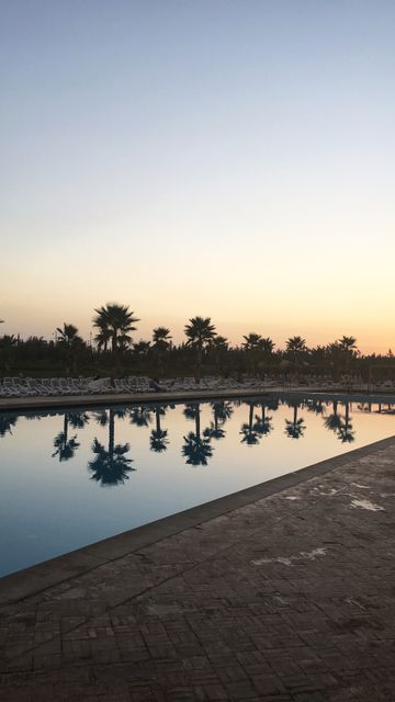 This image captures a tranquil evening scene with the reflections of palm trees in a serene swimming pool during sunset. It is perfect for use in travel brochures, vacation advertisements, wellness and spa promotions, or any content that evokes relaxation and calm.